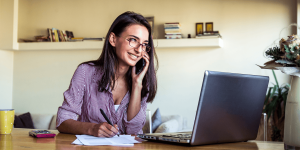 Mulher trabalhando em home office, sorrindo enquanto fala ao telefone, com notebook, papéis e caneta sobre a mesa, representando um ambiente de trabalho remoto.